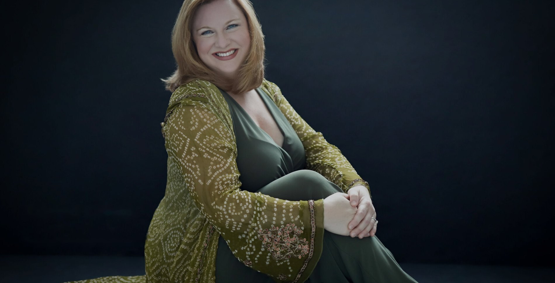 Kristen Calvin seated against a dark backdrop in an olive jacket, smiling